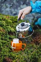 The hand holds the kettle in nature to pour boiling water into a mug, brew a coffee filter bag, an instant drink in nature, a quick preparation of breakfast. photo