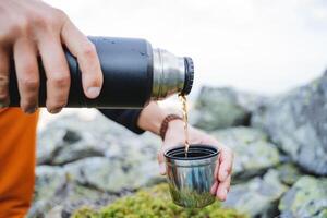 The hand holds a thermos, water flows from the flask to the mug, a hot drink to drink in nature, coffee in the morning in the mountains, hands to hold a glass of tea, a tourist drinks a hot drink. photo