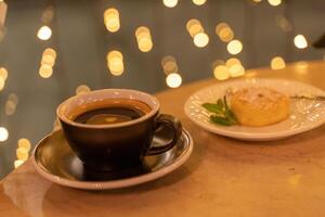 A cup of coffee sits on a table next to a plate of dessert. The dessert is a small cake with a leaf on top. Scene is cozy and inviting, as the coffee and dessert are perfect for a relaxing evening. photo