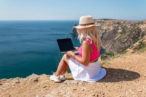 A woman is sitting on a rocky beach with a laptop open in front of her. She is wearing a pink top and a straw hat. Concept of relaxation and leisure. photo