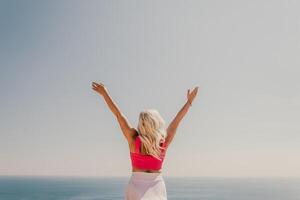 A woman in a pink top and white skirt is standing on a cliff overlooking the ocean. She is smiling and she is happy. photo