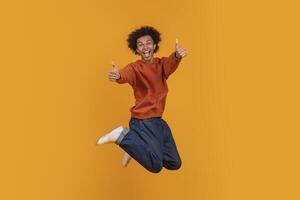 African American young man with an afro is jumping in the air with both thumbs up. He is wearing a red sweater and blue jeans. He is smiling and looking directly at the camera photo
