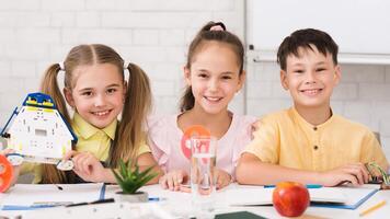 Three children, a girl holding a robot and two others, are sitting at a table in a classroom setting, smiling and working on a STEM project. photo