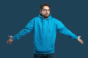 A young man stands with outstretched arms, indicating confusion or disbelief while wearing a bright blue hoodie. The background color drawing attention to his expression and attire. photo
