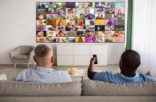 Two men are relaxing on a couch in a living room. They are watching television. The television screen is displaying a collage of many different images. One man is holding a remote control. photo