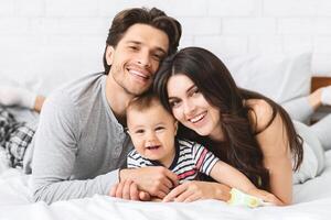 A smiling family of three lies on a bed together. The parents are both looking at the camera, while the baby is smiling at someone out of frame. The family looks relaxed and happy. photo
