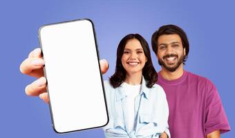 A young multiethnic couple is smiling and holding a smartphone with a blank screen. The man is wearing a purple t-shirt and the woman is wearing a light blue shirt. The background is a solid purple photo
