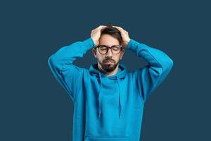 A young man in a blue hoodie displays signs of stress as he holds his head with both hands. The background is a solid color, emphasizing his expression of worry and concern. photo