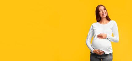 Pregnancy. Cheerful Pregnant Lady Touching Belly Smiling At Camera Standing On Pink Background. Studio Shot photo