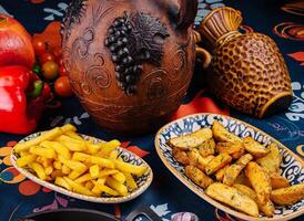 Serving plates overflowing with french fries and potato wedges surrounded by earthenware jugs and fresh vegetables photo