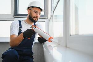 Young man, dressed in a uniform, doing repair in the house. Handyman is applying silicone from a caulk gun to prevent water leak photo