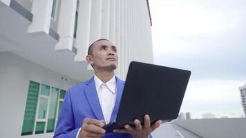 A businessman in a blue suit holds a laptop and looks upward while standing outside a modern building. The scene suggests forward-thinking, technology, and corporate ambition. video