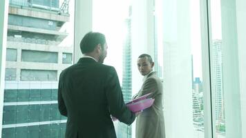 A businessman holding folders approaches a colleague who is standing by a large window, gazing out at the city skyline. The scene conveys reflection and corporate planning. video