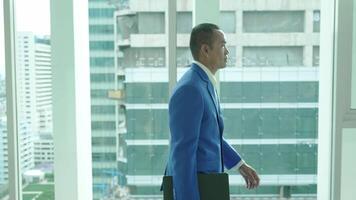 A businessman in a blue suit works on his tablet near a window overlooking the city. Another professional walks by, highlighting a dynamic office environment. video