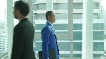 A businessman in a blue suit works on his tablet near a window overlooking the city. Another professional walks by, highlighting a dynamic office environment. video