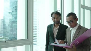 Two business professionals stand by a large window in an office, reviewing documents in a folder. The cityscape in the background reflects a corporate and urban environment. video