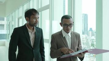 Two businessmen stand by a large office window, reviewing documents in a binder. One man gestures as he explains while the other listens closely, signifying collaboration and teamwork. video