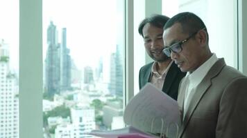 Two business professionals stand by a large window in an office, reviewing documents in a folder. The cityscape in the background reflects a corporate and urban environment. video