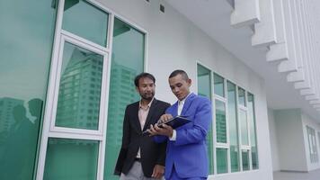 Two businessmen stand outside a modern office building, reviewing information on a laptop. One man explains something while the other listens attentively, reflecting teamwork and collaboration. video