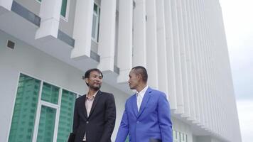Two businessmen in formal suits walk together outside a modern building, carrying folders. The sleek architecture in the background suggests a professional or corporate setting. video