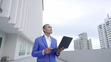 A businessman in a blue suit holds a laptop and looks upward while standing outside a modern building. The scene suggests forward-thinking, technology, and corporate ambition. video