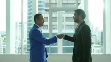 Two businessmen shake hands in an office setting with large windows and a city view. The handshake signifies agreement, partnership, or the conclusion of a successful meeting. video