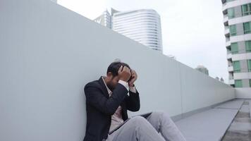 A businessman in a suit sits on the ground outside a modern building, holding his head in frustration. The scene reflects stress, anxiety, or challenges in a corporate environment. video