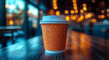 A coffee cup sits on a table in a cafe. The cup is made of paper and has a white lid. The scene is set in a dimly lit room with a blurry background, giving the impression of a cozy photo