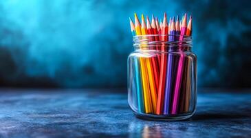 A jar full of colored pencils on a table. The pencils are of different colors and are arranged in a way that they are visible from all angles. The jar is placed on a blue surface photo