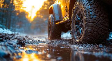A dirty jeep tire is in the mud. The tire is covered in mud and snow, and the ground is wet. The scene is dark and moody, with the sun setting in the background photo