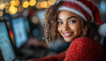 A woman with curly hair is smiling and wearing a red hat and a red sweater. She is sitting in front of a computer with a keyboard and a mouse photo