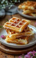 Three waffles stacked on top of each other on a plate. The plate is on a wooden table. There are flowers in the background photo