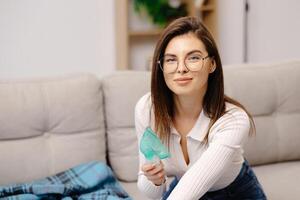 Young woman holding a mask nebulizer inhaling fumes medication into lungs. Caucasian sick lady inhaling through inhaler mask at home. Self treatment of the respiratory tract using inhalation nebulizer photo