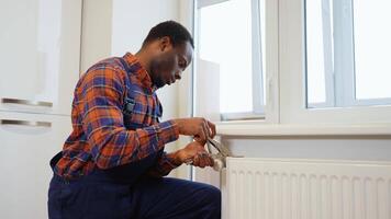 Young black plumber installing heating radiator in an empty room of a newly built apartment or house video