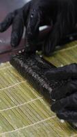 Hands of a professional sushi chef laying out rice on Nori leaves lying on a bamboo mat on a white kitchen table with a row of fresh vegetables, bell peppers and lettuce leaves laid out. video