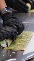 Hands of a professional sushi chef laying out rice on Nori leaves lying on a bamboo mat on a white kitchen table with a row of fresh vegetables, bell peppers and lettuce leaves laid out. video
