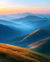 Landscape of majestic mountain ranges at sunrise, with golden sunlight casting soft rays over distant hills and valleys. The foreground consists of a grassy ridge covered in golden autumn hues. photo