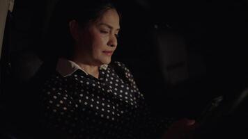 Asian woman sitting in dark car using phone at night. Using smartphone, reading text message on mobile with slight smile. Darkness, driving and stopping at traffic light. Chat message. video