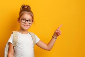 Portrait of cute little girl with glasses and backpack pointing at something on yellow background photo