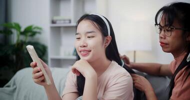Handheld shot, Asian mother combs her daughter's hair while she looks at mobile phone on the sofa in the living room at home. Happy family moment. video
