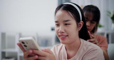 Handheld selective focus shot, Asian mother combs her daughter's hair while she looks at mobile phone on the sofa in the living room at home. Happy family moment. video