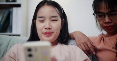 Handheld selective focus shot, Asian mother and young woman are looking at a smartphone together on sofa. They sit closely, sharing a quiet moment as they enjoy something on the screen video