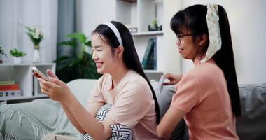 Handheld selective focus shot, Asian mother combs her daughter's hair while she looks at mobile phone on the sofa in the living room at home. Happy family moment. video
