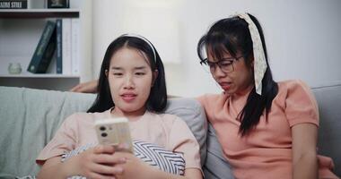 Handheld shot, Asian mother and young woman are looking at a smartphone together on sofa. They sit closely, sharing a quiet moment as they enjoy something on the screen video