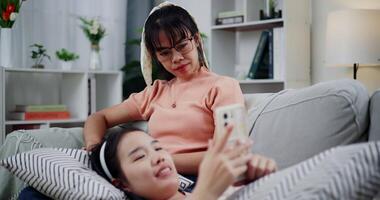 Handheld shot, Daughter is lying on her mother's lap and they are watching something together on a mobile phone on the sofa in the living room, enjoying a fun time together. video