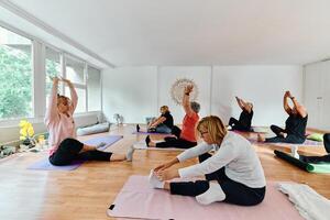 A group of senior women engage in various yoga exercises, including neck, back, and leg stretches, under the guidance of a trainer in a sunlit space, promoting well-being and harmony photo