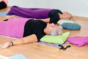 A group of senior women engage in various yoga exercises, including neck, back, and leg stretches, under the guidance of a trainer in a sunlit space, promoting well-being and harmony photo