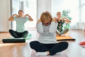 A group of senior women engage in various yoga exercises, including neck, back, and leg stretches, under the guidance of a trainer in a sunlit space, promoting well-being and harmony photo
