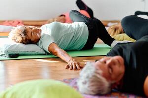 A group of senior women engage in various yoga exercises, including neck, back, and leg stretches, under the guidance of a trainer in a sunlit space, promoting well-being and harmony photo