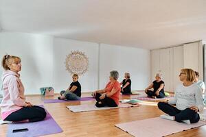A group of senior women engage in various yoga exercises, including neck, back, and leg stretches, under the guidance of a trainer in a sunlit space, promoting well-being and harmony photo